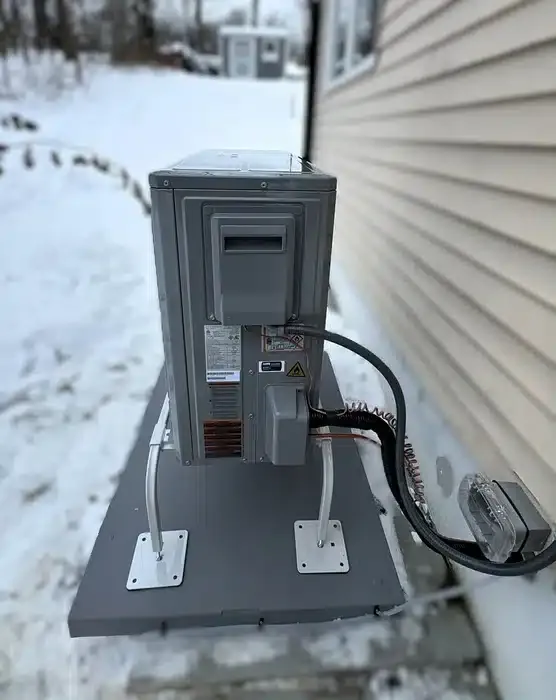Outdoor heat pump condensing unit mounted on a raised stand beside a Newfoundland, NJ home in winter