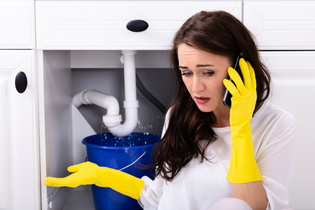 Sad Young Woman Calling Plumber In Front Of Water Leaking From Sink Pipe