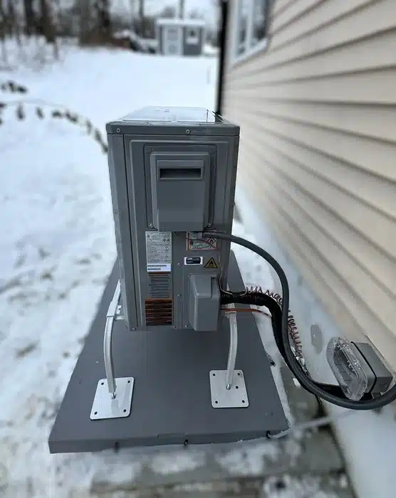 Outdoor heat pump condensing unit mounted on a raised stand beside a Newfoundland, NJ home in winter